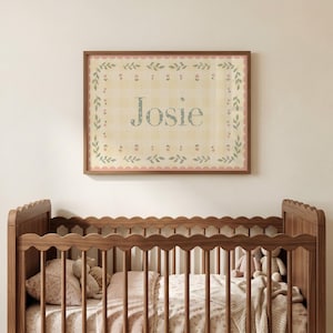 May include: A rectangular wall art print in a wooden frame featuring the name Josie in blue letters. The background has a yellow gingham pattern with a border of green leaves and small pink flowers, finished with a scalloped pink edge.