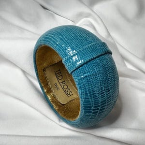 May include: A turquoise blue bangle bracelet with a textured, reptile-like pattern. The interior is lined with gold fabric. A rectangular tag with the text "TED ROSSI NYC" is visible.