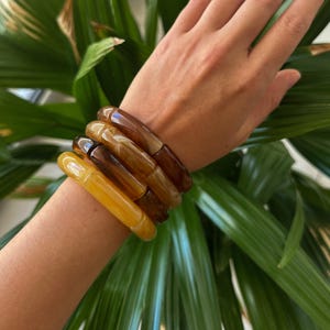 Op de afbeelding: Een stapel van vijf dikke, doorschijnende armbanden in tinten amber, honing en bruin. De cilindrische armbanden worden om een pols gedragen, met groene bladeren op de achtergrond.