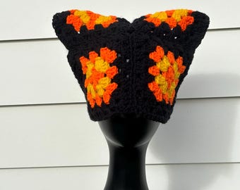 May include: A black crochet hat with orange and yellow floral patterns. The hat is displayed on a black mannequin head against a white background. The hat's design features square motifs.