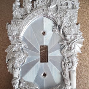 May include: Silver-colored light switch cover with a fantasy theme. The cover features detailed relief sculptures of dragons, castles, and shields. The central area has a geometric pattern around the switch opening.