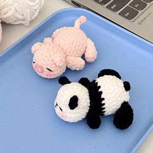 May include: Two crocheted stuffed animals: a pink pig and a black and white panda. The pig has a small tail and closed eyes. The panda has black limbs and eye patches. Both are lying on a light blue tray, near a laptop.