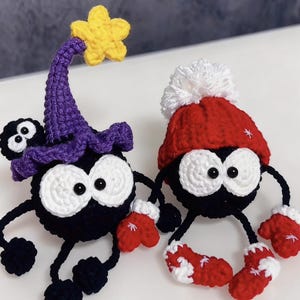 May include: Two handmade crochet figures. One is black with a purple witch hat and yellow star. The other is black with a red hat and mittens. Both have large white eyes and red and white accents.
