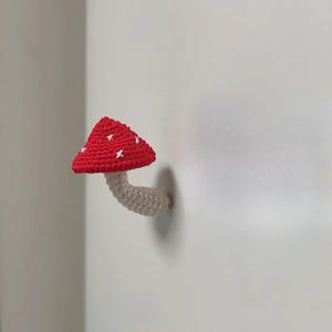 May include: A crocheted mushroom with a red cap and white spots is mounted on a white surface. The stem is beige. This handmade item is a decorative piece, possibly a wall hanging or ornament.