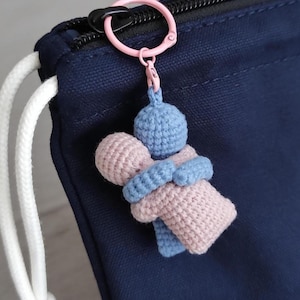 Può includere: Un portachiavi all'uncinetto fatto a mano con due figure che si abbracciano. Le figure sono in azzurro e rosa, collegate da un abbraccio blu. Il portachiavi ha un anello rosa ed è attaccato a una borsa blu navy con un cordoncino bianco.