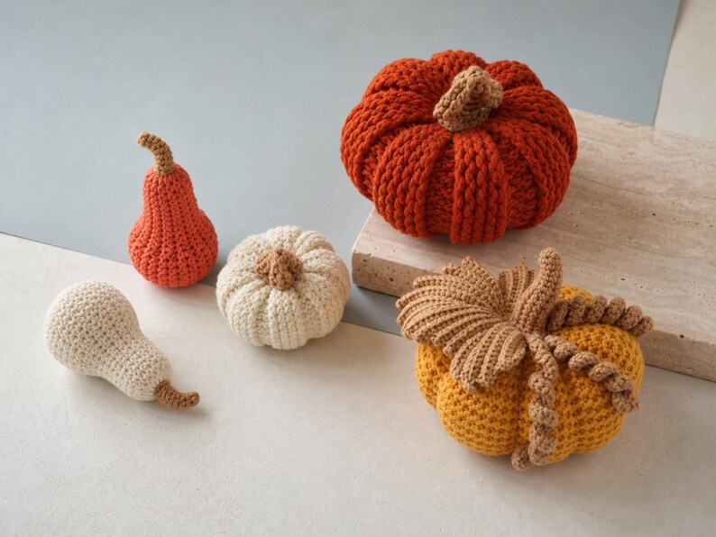 May include: A collection of five handmade crochet items including a large orange pumpkin, a smaller cream pumpkin, a small orange gourd, a small cream pear-shaped gourd, and a yellow pumpkin decorated with brown crocheted leaves and vines.