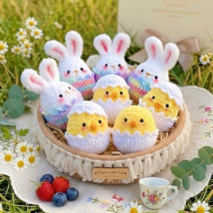May include: A decorative basket contains crocheted Easter bunnies and chicks. The bunnies have white bodies, pink ears, and rainbow-striped sweaters. The chicks are yellow and in white crocheted eggshells. A small teacup and berries are also present.