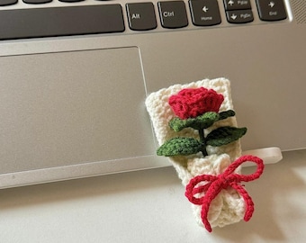 May include: A handmade crochet bouquet featuring a red rose with green leaves and a white crocheted wrap. The bouquet is tied with a red bow. The item is placed on a laptop.