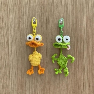 May include: Two handmade crochet keychains: a yellow duck with orange accents and a green frog with a pink tongue. Both have large googly eyes and yellow or green metal clasps. The keychains are displayed against a light brown wooden background.