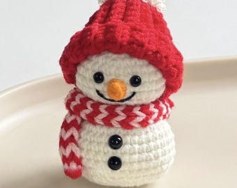 May include: A crocheted snowman figure. The snowman is white with a red hat and a white bobble, and a red and white scarf. It has a carrot nose, black button eyes, and black button details. A festive decoration.
