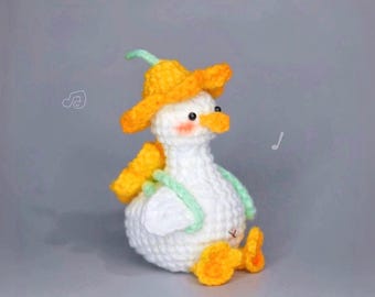 May include: A handmade crochet duckling wearing a yellow hat and holding a yellow flower. The duckling is white with an orange beak and feet. The duckling has a green strap and is sitting down. The background is a solid gray.