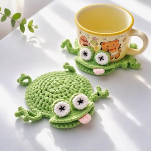 May include: Two handmade green crochet frog coasters with white and black eyes and pink tongues. One coaster has a yellow mug with a bear design on top. The coasters are on a white surface.