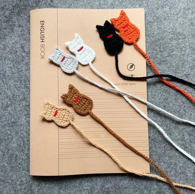 May include: Crocheted cat bookmarks in various colors, including white, gray, black, orange, and brown, each with a red accent. The bookmarks are placed on a notebook with the text "ENGLISH BOOK".