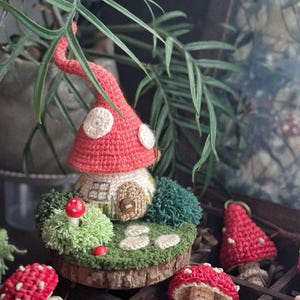 May include: A crocheted mushroom house with a red cap and white spots, set on a wooden base with green moss and small red mushrooms. Other crocheted mushrooms are in the background.