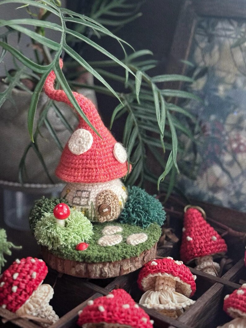 May include: A crocheted mushroom house with a red cap and white spots, set on a wooden base with green moss and small red mushrooms. Other crocheted mushrooms are in the background.