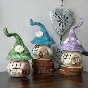 May include: Three whimsical crochet mushroom houses in green, blue, and purple, each with a white door and window. The houses sit on wooden slices, showcasing a handcrafted, fairy-tale aesthetic. A decorative heart hangs in the background.