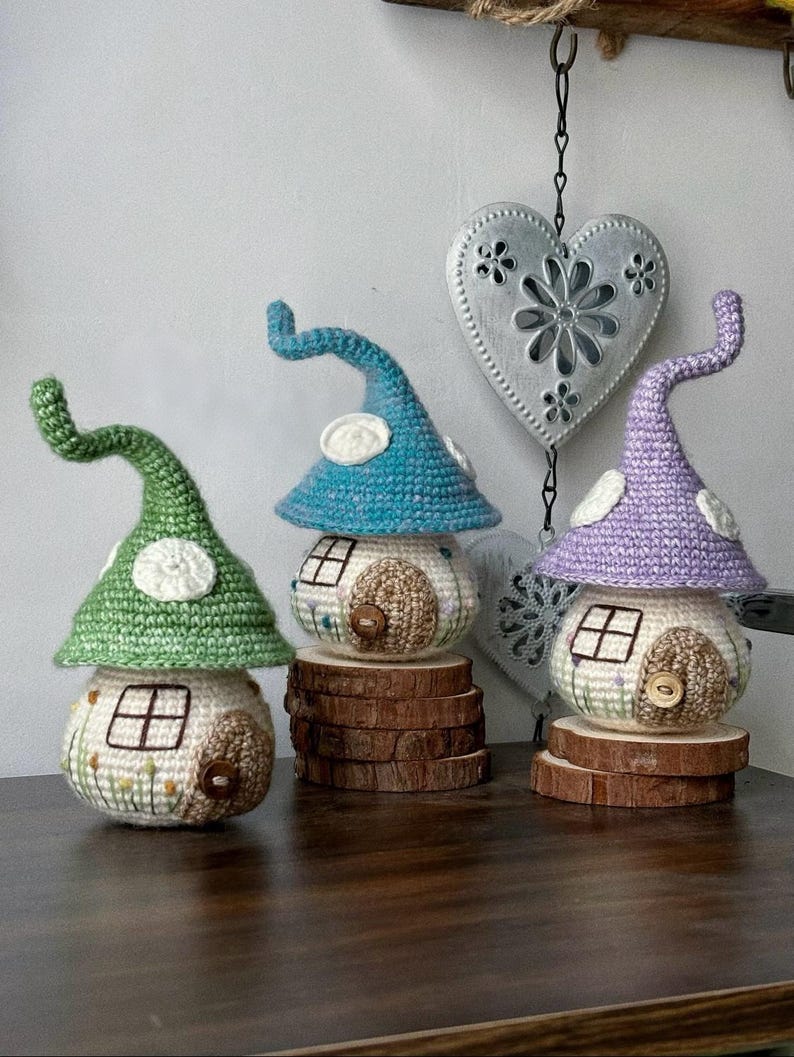 May include: Three colourful crochet mushroom houses in green, blue, and purple, each with a white door and window. The houses sit on wooden slices, showcasing a handcrafted, fairy-tale aesthetic. A decorative heart hangs in the background.
