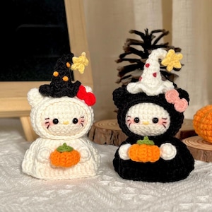 May include: Two crochet figures, one white and one black, each resembling a cat. The white figure wears a witch hat with orange accents and holds an orange pumpkin. The black figure wears a ghost hat and holds a pumpkin. Halloween-themed decor.