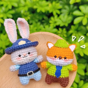 May include: Two handmade crochet amigurumi figures: a bunny wearing a blue hat and uniform, and a fox in a green jacket and tie. The bunny has pink cheeks and the fox has an orange head. Both are on a wooden surface.