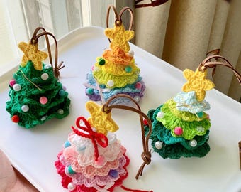 May include: Four handmade crochet Christmas tree ornaments in various colors. Each tree has a yellow star on top and a brown leather loop for hanging. The trees are decorated with beads and bows, and are displayed on a white tray.