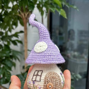 May include: A handmade crochet mushroom house. It features a lavender cap with white spots and a beige body with a brown door, a small window, and embroidered flowers. A button is the door handle.