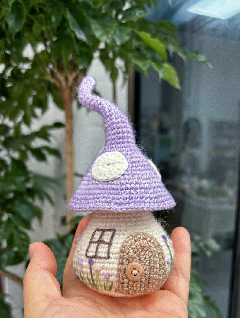 May include: A handmade crochet mushroom house. It features a lavender cap with white spots and a beige body with a brown door, a small window, and embroidered flowers. A button is the door handle.