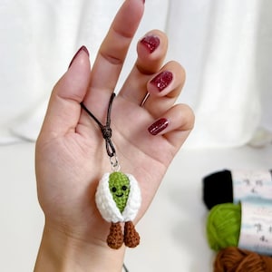 Peut inclure: Pendentif fait main au crochet représentant un personnage vert souriant, avec une écharpe blanche et des pieds marron, suspendu à un cordon marron. L'arrière-plan comprend des pelotes de laine.