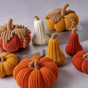 May include: A collection of eight crocheted pumpkins and gourds in various sizes and shapes. The items feature textured yarn in shades of orange, yellow, and cream, with brown stems and decorative crocheted leaves on the larger round pumpkins.