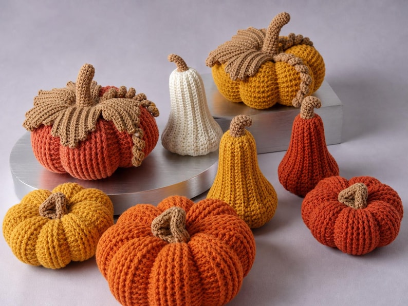May include: A collection of eight crocheted pumpkins and gourds in various sizes and shapes. The items feature textured yarn in shades of orange, yellow, and cream, with brown stems and decorative crocheted leaves on the larger round pumpkins.