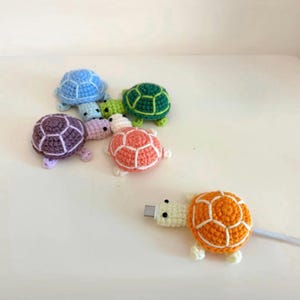 May include: A collection of colorful, crocheted turtle-shaped cable protectors. The turtles are in various colors, including blue, green, purple, pink, and orange. The orange turtle is attached to a USB cable. The turtles have white accents and are arranged on a white surface.