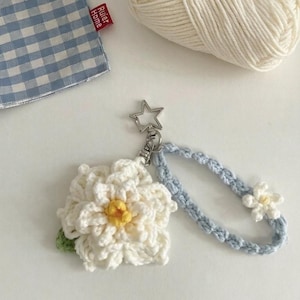 May include: A handmade crochet keychain featuring a large white flower with a yellow centre and a small green leaf. The keychain includes a light blue braided wrist strap and a small white flower. A silver star-shaped clasp is attached.