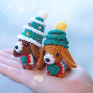 May include: Two crocheted dachshund dog ornaments, each wearing a Christmas tree hat. The dogs are brown and white with green, red, and white accents. One hat has a white ball on top, the other a yellow star.