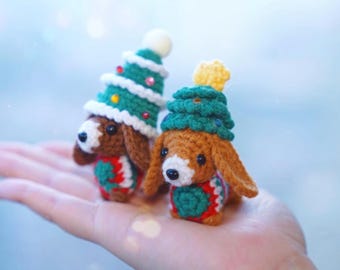 以下が含まれることがあります： クリスマスツリーの帽子をかぶった、2匹のかぎ針編みのダックスフンド犬のオーナメント。犬は茶色と白で、緑、赤、白のアクセントが付いています。片方の帽子には白いボール、もう片方には黄色の星が付いています。