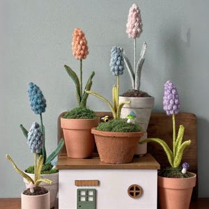 Puede incluir: Una colección de flores de ganchillo hechas a mano en macetas de terracota y una casa en miniatura. Las flores son en tonos de azul, rosa, naranja y morado. La casa tiene una puerta verde y una ventana redonda.