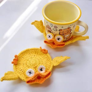 May include: Two handmade yellow crochet duck coasters with orange beaks and white eyes with black crosses. One coaster holds a yellow mug with a bear and food design. The coasters are on a white surface.