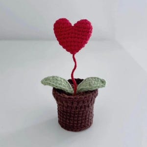 May include: A handmade crochet plant featuring a red heart-shaped flower, green leaves, and a brown pot. The heart flower has a long red stem. The pot is a dark brown colour.