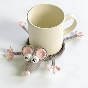 Könnte beinhalten: Ein cremefarbener Becher ruht auf einem grau gehäkelten mausförmigen Untersetzer. Der Untersetzer hat rosa Ohren, Pfoten und eine Nase mit weißen Augen und schwarzen Kreuzdetails. Der Becher steht auf einer weißen Oberfläche.