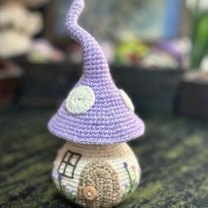 May include: A handmade crochet mushroom house. It features a lavender cap with white spots and a beige base with a door, window, and floral embroidery. A charming decorative item.