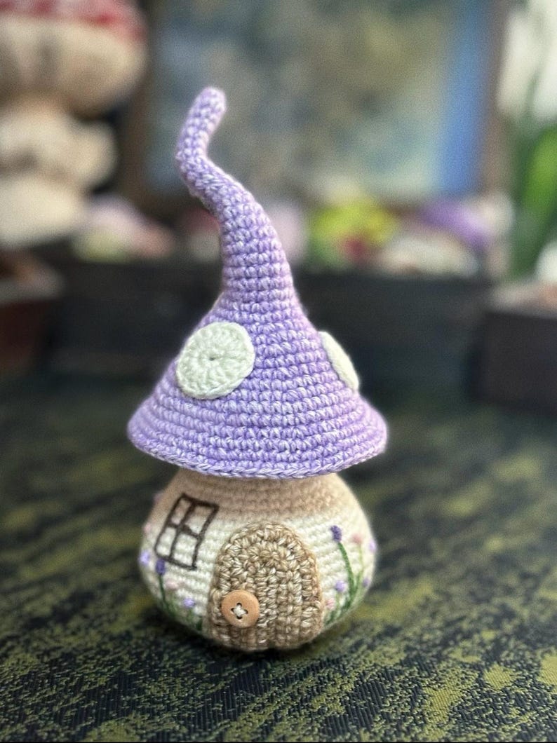 May include: A handmade crochet mushroom house. It features a lavender cap with white spots and a beige base with a door, window, and floral embroidery. A charming decorative item.