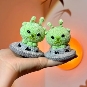 May include: Two handmade, green alien plush toys with black eyes and antennae, sitting on gray UFO-shaped bases. The plushies are held in a hand, showcasing the detail of the crochet work. The background is a warm, soft orange.