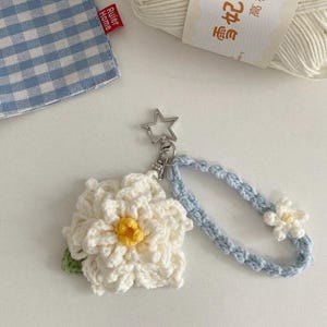 Puede incluir: Un llavero de flores hecho a mano con un diseño de margarita blanca, centro amarillo y hoja verde. El llavero tiene una correa de muñeca azul claro y un cierre plateado en forma de estrella. Un tejido a cuadros azul y blanco y un ovillo de lana están en el fondo.