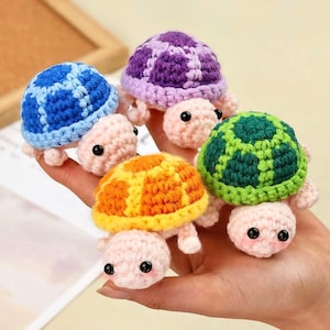 May include: Five colourful, handmade crochet turtles. The shells are blue, purple, green, orange, and yellow. Each turtle has a textured shell and a light pink head, legs, and tail. They have black eyes and are held in a hand.