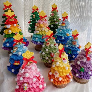 Puede incluir: Una colección de árboles de Navidad de ganchillo coloridos. Cada árbol tiene una estrella en la parte superior, adornos decorativos y está montado sobre una base de madera. Los árboles son de varios colores, incluyendo verde, azul, amarillo y rosa.