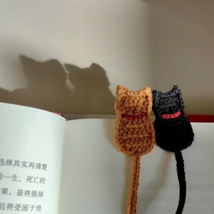 May include: Two handmade crochet cat bookmarks. One is brown with a red collar, the other is black with a red collar. The bookmarks are placed in an open book with Chinese text. The cat bookmarks are a unique gift for book lovers.