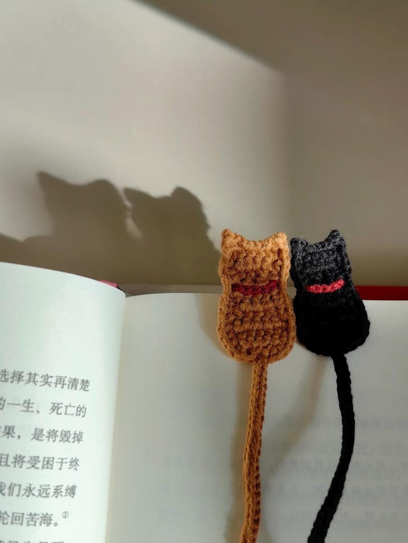 May include: Two handmade crochet cat bookmarks. One is brown with a red collar, the other is black with a red collar. The bookmarks are placed in an open book with Chinese text. The cat bookmarks are a unique gift for book lovers.