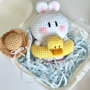 May include: A handmade crochet bunny with pink cheeks and a yellow duck floatie. A small crochet sun hat with a white bow sits next to the bunny. The items are presented in a white container with blue paper filler.