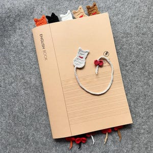 May include: A tan notebook with the words "ENGLISH BOOK" printed on the cover. Several crocheted cat bookmarks in orange, black, white, tan, and brown are inserted into the top of the notebook. A gray cat bookmark with a red bow is attached to the notebook.