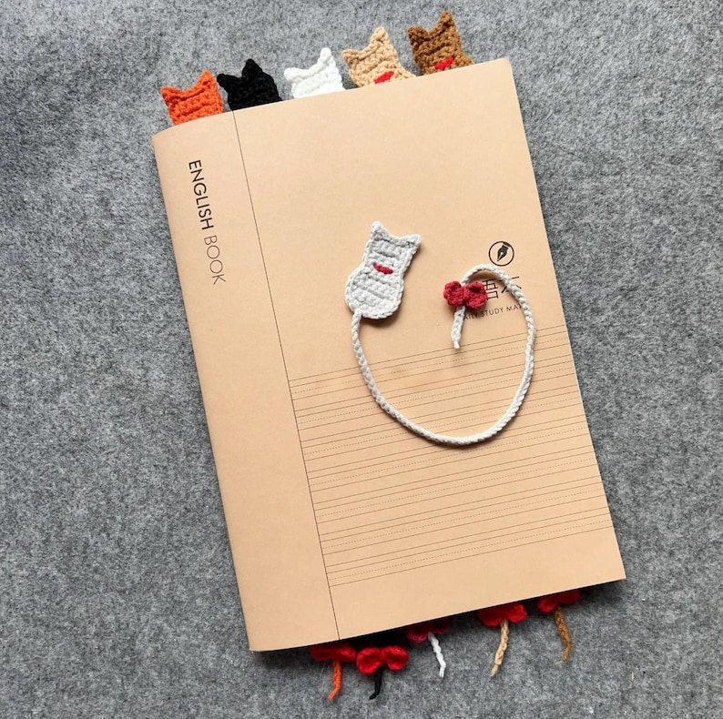May include: A tan notebook with the words "ENGLISH BOOK" printed on the cover. Several crocheted cat bookmarks in orange, black, white, tan, and brown are inserted into the top of the notebook. A gray cat bookmark with a red bow is attached to the notebook.