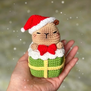 May include: A crocheted capybara wearing a red Santa hat and a red bow tie, inside a green gift box with yellow ribbon. The capybara is tan with black eyes and a black nose. The gift box has a white trim.