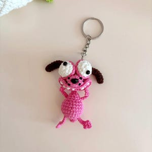 May include: A handmade pink crochet keychain featuring the cartoon character Courage the Cowardly Dog. The keychain has large white eyes, brown ears, and a pink body. The keychain is attached to a silver metal ring.